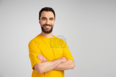 Téléchargez les photos : Heureux homme barbu en t-shirt jaune souriant à la caméra tout en se tenant debout avec les bras croisés isolés sur gris - en image libre de droit