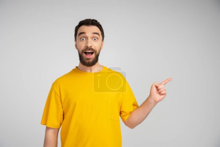 surprised man in yellow t-shirt looking at camera and pointing with finger isolated on grey