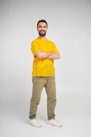 Foto de Longitud completa de hombre barbudo feliz en camiseta amarilla y pantalones beige de pie con brazos cruzados sobre fondo gris - Imagen libre de derechos