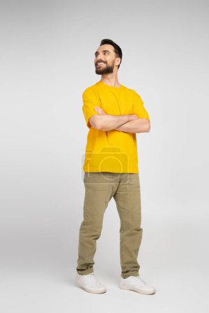 Photo for Full length of happy man in beige pants and yellow t-shirt standing with crossed arms and looking away on grey - Royalty Free Image