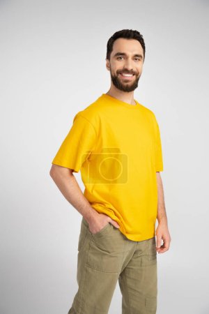 brunette man in yellow t-shirt standing with hand in pocket and smiling at camera isolated on grey