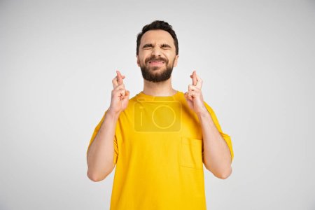 worried man in yellow t-shirt standing with closed eyes and crossed fingers isolated on grey