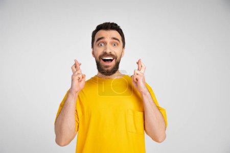nervous man in yellow t-shirt holding crossed fingers for luck isolated on grey