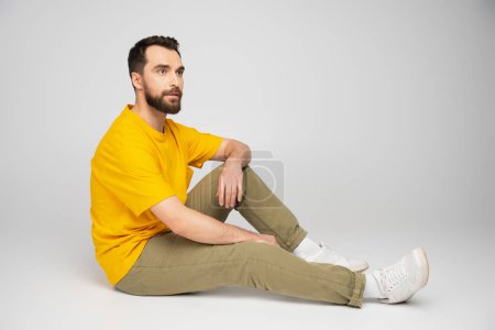 Foto de Longitud completa del hombre en pantalones beige y camiseta amarilla mirando hacia otro lado mientras está sentado sobre fondo gris - Imagen libre de derechos