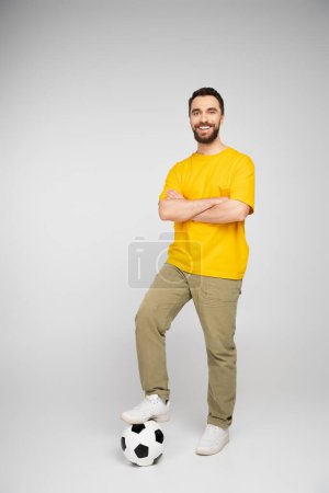 longitud completa del hombre feliz pisando la pelota de fútbol mientras está de pie con los brazos cruzados sobre fondo gris