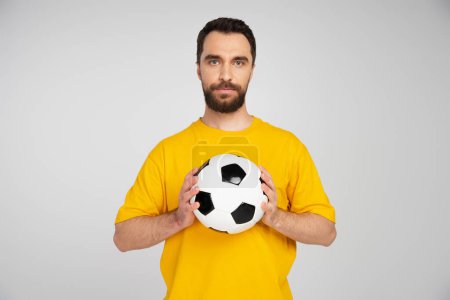 bearded sports fan in yellow t-shirt holding soccer ball while looking at camera  isolated on grey