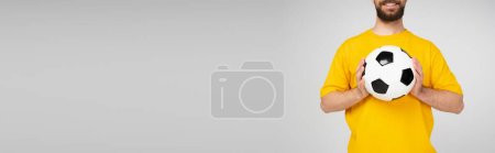 Foto de Vista recortada del hombre barbudo y sonriente en camiseta amarilla sosteniendo pelota de fútbol aislado en gris, bandera - Imagen libre de derechos