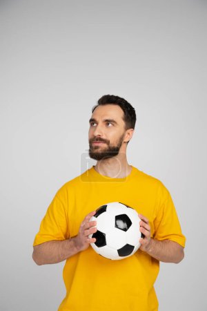 brünetter bärtiger Fußballfan in gelbem T-Shirt, Ball in der Hand und isoliert auf grau blickend