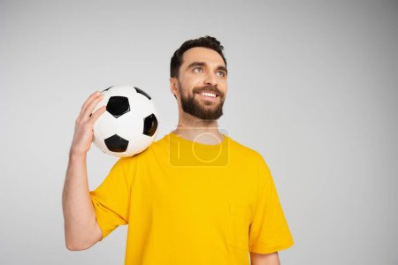 glücklicher bärtiger Sportfan in gelbem T-Shirt, der einen Fußballball in der Hand hält und isoliert auf grau blickt