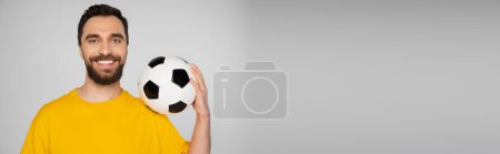cheerful bearded man in yellow t-shirt holding soccer ball isolated on grey, banner