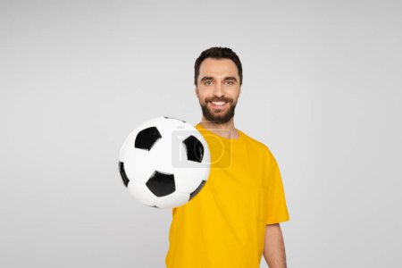 positive football fan in yellow t-shirt showing soccer ball at camera isolated on grey