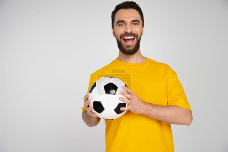 Foto de Fanático del fútbol barbudo excitado en camiseta amarilla sosteniendo la pelota y riéndose de la cámara aislada en gris - Imagen libre de derechos