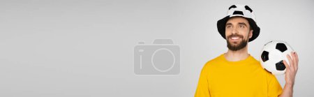 Photo for Happy man in football fan hat standing with soccer ball and looking away isolated on grey, banner - Royalty Free Image