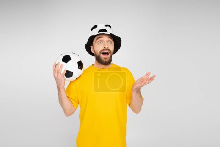 amazed bearded man in football fan hat holding soccer ball and pointing with hand isolated on grey