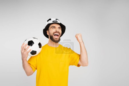 Aufgeregter Mann mit Fußballfanhut hält Fußballball in der Hand und zeigt Triumphgeste isoliert auf grau