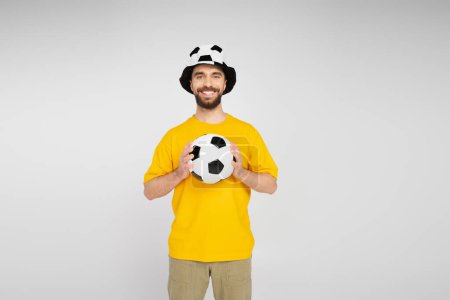 Foto de Alegre barbudo hombre en fútbol abanico sombrero celebración fútbol pelota y mirando cámara aislado en gris - Imagen libre de derechos