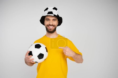 happy man in football fan hat pointing at soccer ball while looking at camera isolated on grey