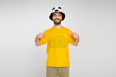 cheerful bearded man in football fan hat and yellow t-shirt pointing with fingers at himself isolated on grey