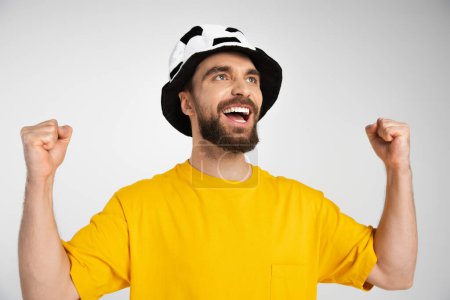 joyful bearded man in football fan hat showing win gesture and shouting isolated on grey