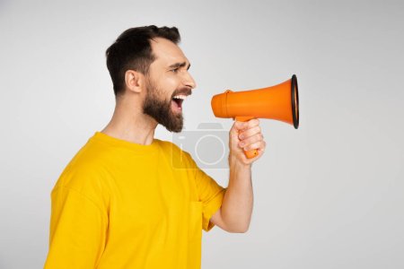 Téléchargez les photos : Vue latérale de l'homme irrité en t-shirt jaune criant en mégaphone isolé sur gris - en image libre de droit