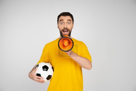 amazed bearded man with soccer ball shouting in megaphone isolated on grey