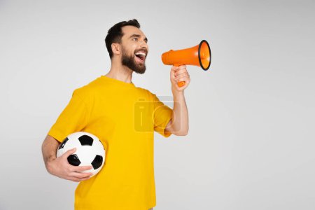 cheerful bearded man shouting in loudspeaker while holding soccer ball isolated on grey