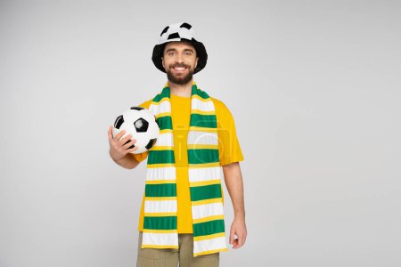 fröhlicher Fußballfan mit Hut und gestreiftem Schal, der Ball in der Hand hält und vereinzelt in die Kamera schaut
