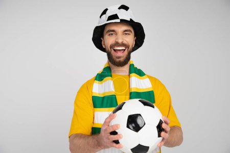 excited man in football fan hat and striped scarf holding ball and laughing at camera isolated on grey
