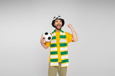 Aufgeregter Sportfan mit Hut und Schal hält Fußballball in der Hand und zeigt Triumphgeste isoliert auf Grau