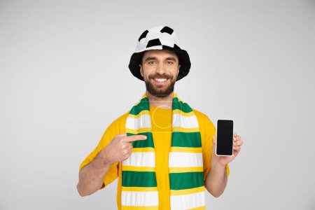 Foto de Hombre barbudo feliz en el sombrero del ventilador de fútbol apuntando al teléfono inteligente con pantalla en blanco aislado en gris - Imagen libre de derechos