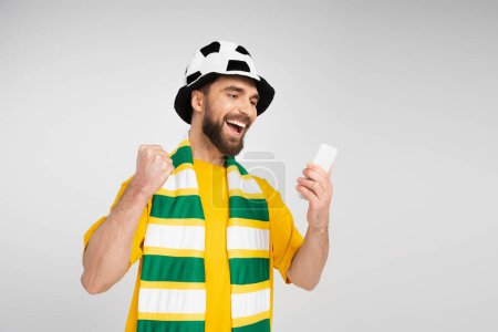 Foto de Hombre emocionado en el sombrero del ventilador de fútbol y bufanda rayada mostrando gesto de victoria mientras ve el partido en el teléfono inteligente aislado en gris - Imagen libre de derechos