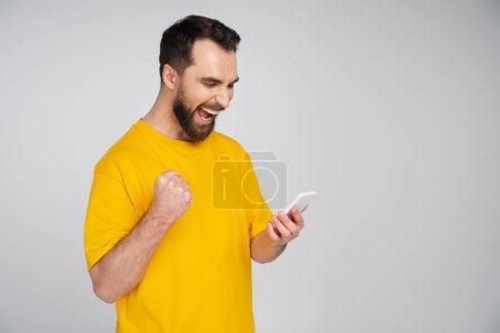 excited bearded man with smartphone showing win gesture and shouting isolated on grey