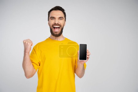 thrilled bearded man holding smartphone with blank screen and showing triumph gesture while screaming isolated on grey