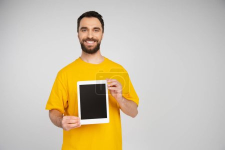 fröhlicher Mann mit Bart zeigt digitales Tablet mit leerem Bildschirm, während er in die Kamera lächelt, isoliert auf grau