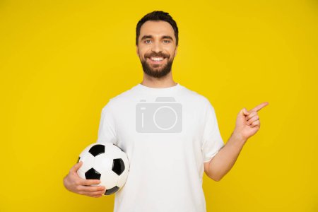 bearded sports fan in white t-shirt holding soccer ball and pointing with finger isolated on yellow
