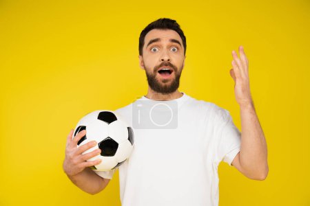 Geschockter und frustrierter Sportfan hält Fußballball in der Hand und gestikuliert isoliert auf gelb
