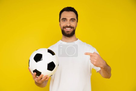 joyful brunette man in white t-shirt pointing at soccer ball isolated on yellow