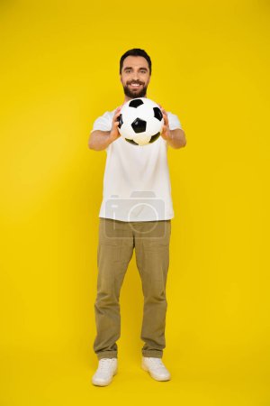 full length of pleased man in pants holding soccer ball in outstretched hands on yellow background