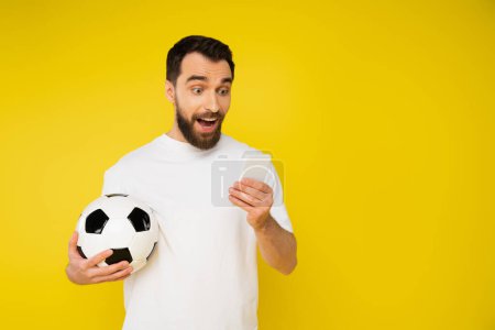 amazed and happy sports fan with soccer ball watching football match on cellphone isolated on yellow