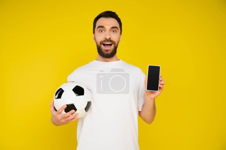 amazed football fan holding ball and smartphone with blank screen isolated on yellow