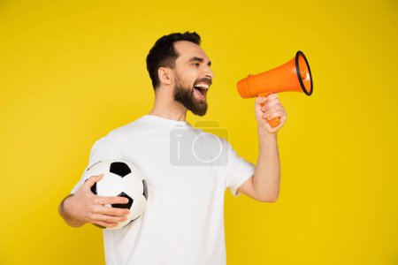 Foto de Abanico de deportes barbudos en camiseta blanca sosteniendo pelota de fútbol y gritando en megáfono aislado en amarillo - Imagen libre de derechos