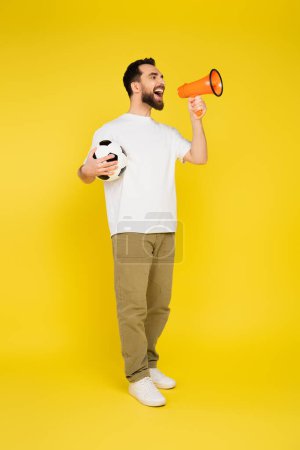 full length of man in white t-shirt and beige pants holding soccer ball and screaming in megaphone on yellow background