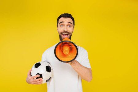 Foto de Admirador de los deportes sorprendidos con pelota de fútbol gritando en megáfono y mirando a la cámara aislada en amarillo - Imagen libre de derechos
