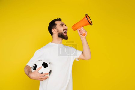excited football fan shouting in loudspeaker while holding ball isolated on yellow