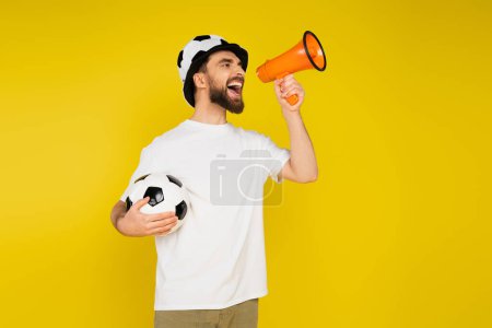 thrilled sports fan in hat standing with soccer ball while shouting in loudspeaker isolated on yellow