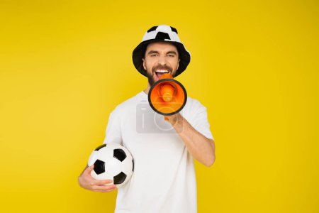thrilled man in football fan hat standing with soccer ball and shouting in loudspeaker isolated on yellow