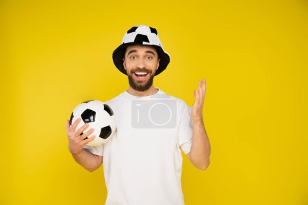Foto de Hombre barbudo feliz en el ventilador de fútbol sombrero sosteniendo pelota de fútbol y mirando a la cámara aislada en amarillo - Imagen libre de derechos