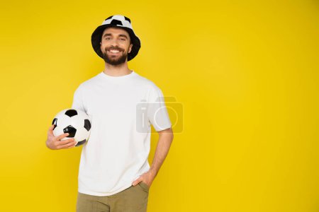 Photo for Cheerful football fan in hat holding ball and standing with hand in pocket isolated on yellow - Royalty Free Image