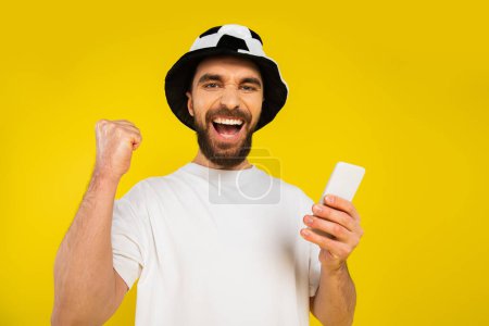 cheerful football fan in hat showing triumph gesture and holding cellphone isolated on yellow