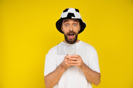 astonished man in football fan hat holding mobile phone and looking at camera isolated on yellow